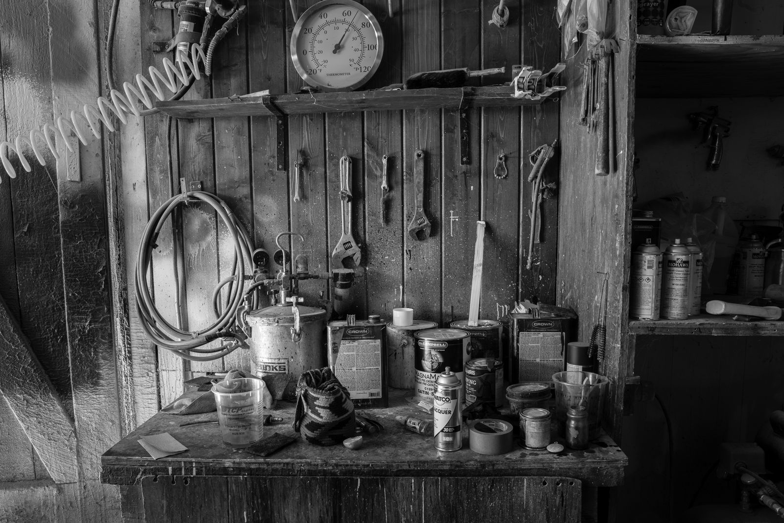The finishing bench beneath a plank wall hung with clamps, coiled hose and hand tools, its top covered in lacquer and stain cans.