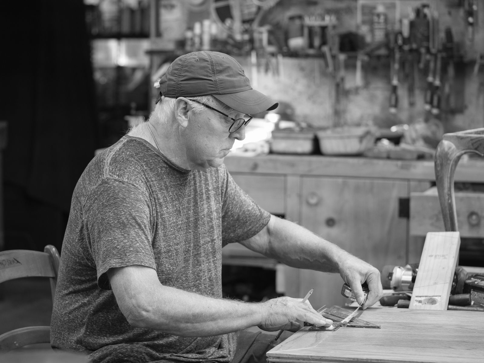 A craftsman in a cap and glasses working a piece by hand at the bench, a curved chair leg clamped upright beside him.