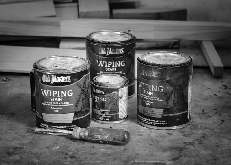 Four cans of Old Masters wiping stain grouped on a bench beside a screwdriver.