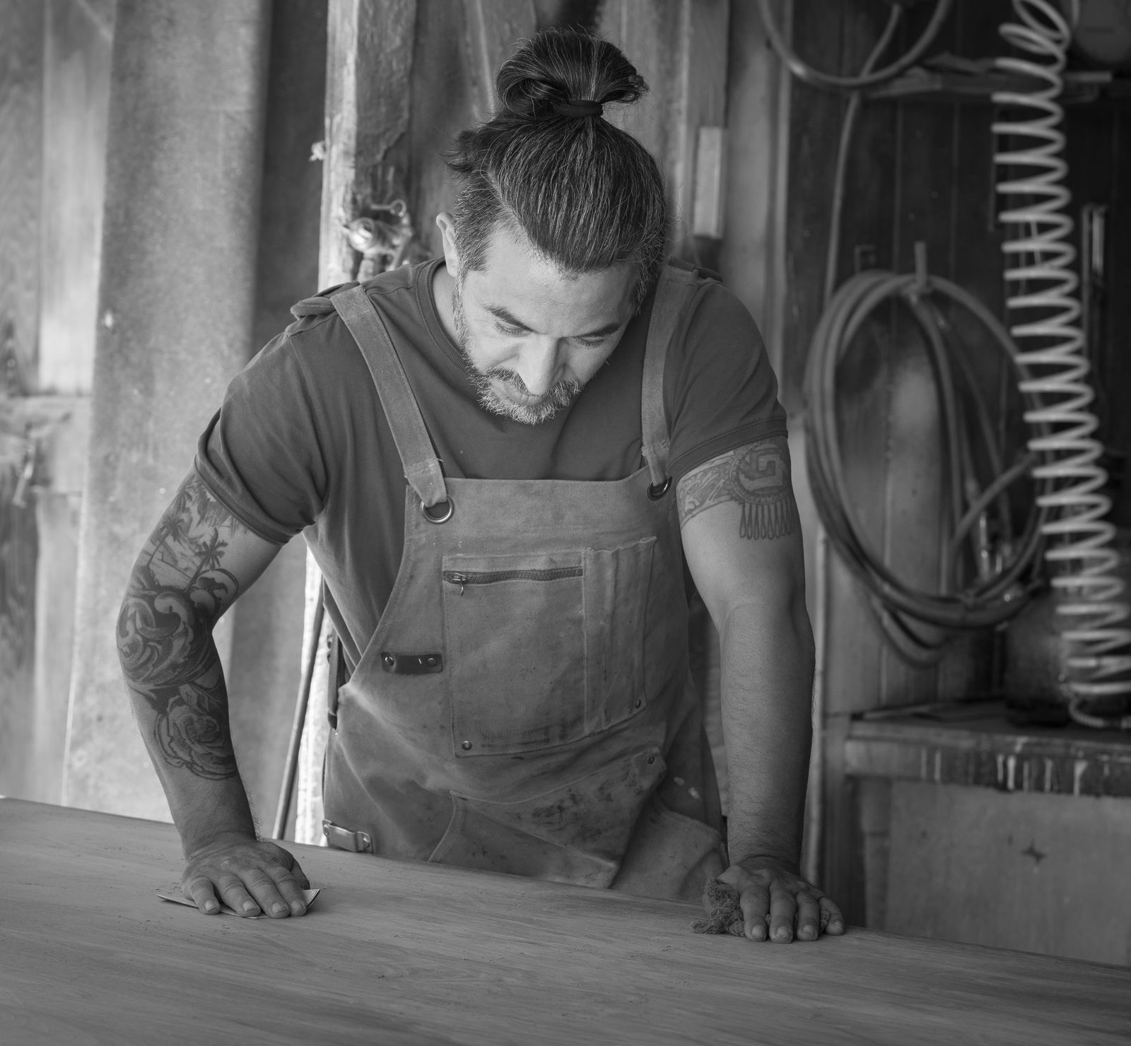 A woodworker in a canvas apron leaning over a tabletop, sighting along the surface with both hands resting on it.