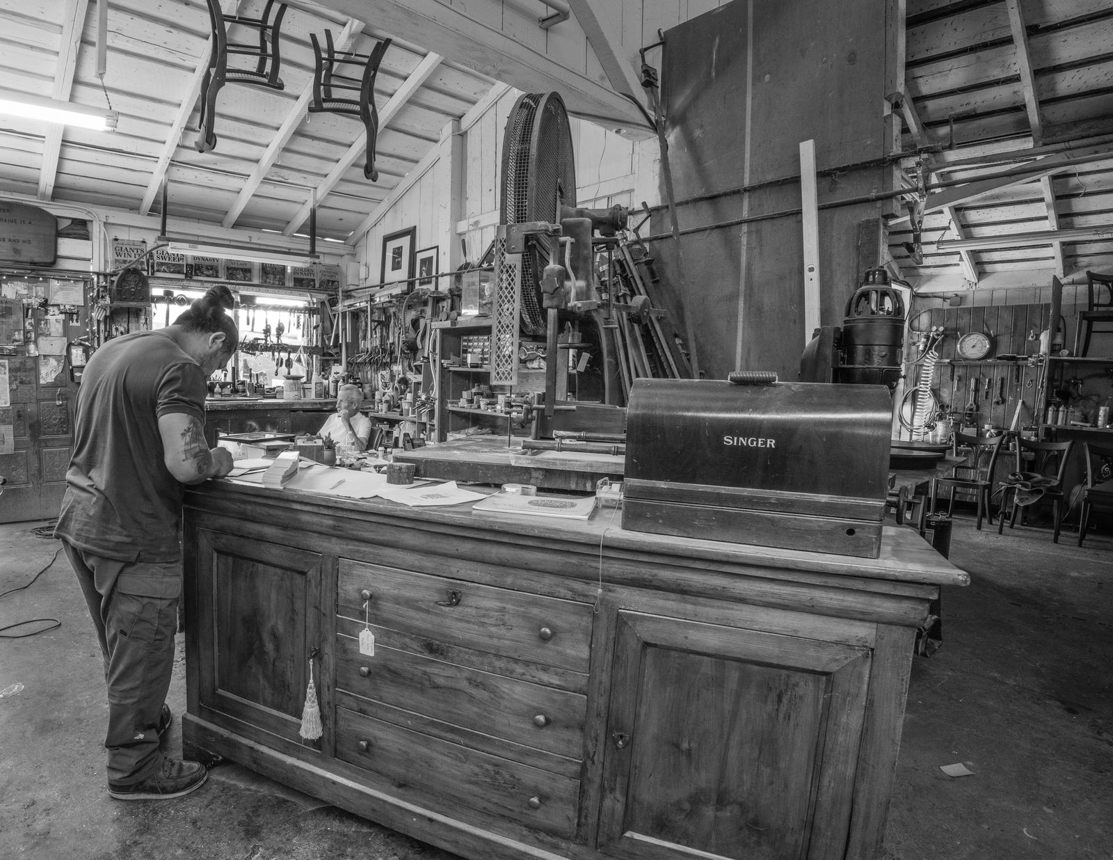 A craftsman at work on a sideboard in the middle of the barn workshop, benches and racked chairs running back behind him.